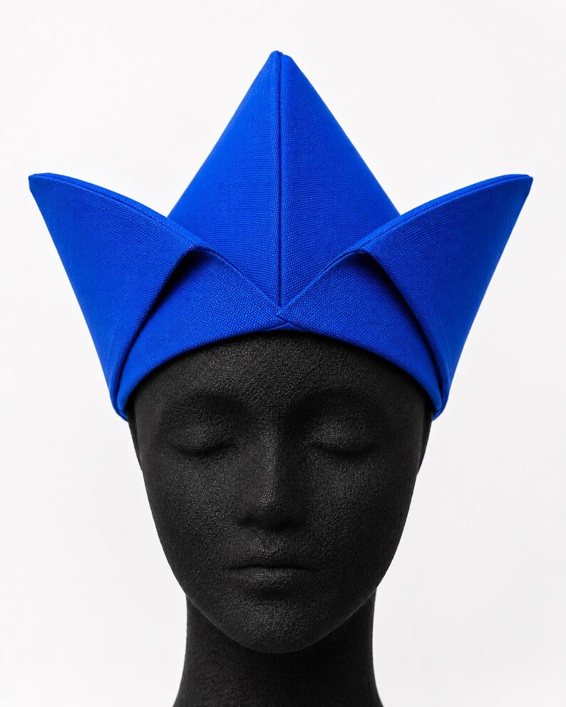 May include: A vibrant blue hat with a distinctive, angular design. The hat features a central, pointed crown and four outward-facing, triangular flaps. The hat is made of a smooth, solid-colored fabric and is displayed on a black mannequin head.