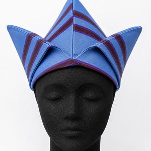 May include: A blue and burgundy striped hat with a unique, pointed design. The hat is displayed on a black mannequin head, showcasing its shape and details. The hat's fabric has a subtle sheen, adding to its visual appeal.