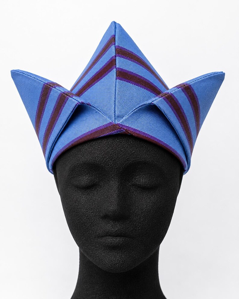 May include: A blue and burgundy striped hat with a unique, pointed design. The hat is displayed on a black mannequin head, showcasing its shape and details. The hat's fabric has a subtle sheen, adding to its visual appeal.