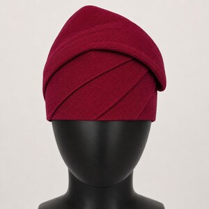 Peut inclure: Un chapeau bordeaux au design superpos&eacute; unique. Le chapeau est fait d'un tissu c&ocirc;tel&eacute;, avec des plis horizontaux cr&eacute;ant un aspect textur&eacute;. Il est pr&eacute;sent&eacute; sur une t&ecirc;te de mannequin noire, mettant en valeur sa forme et son style.