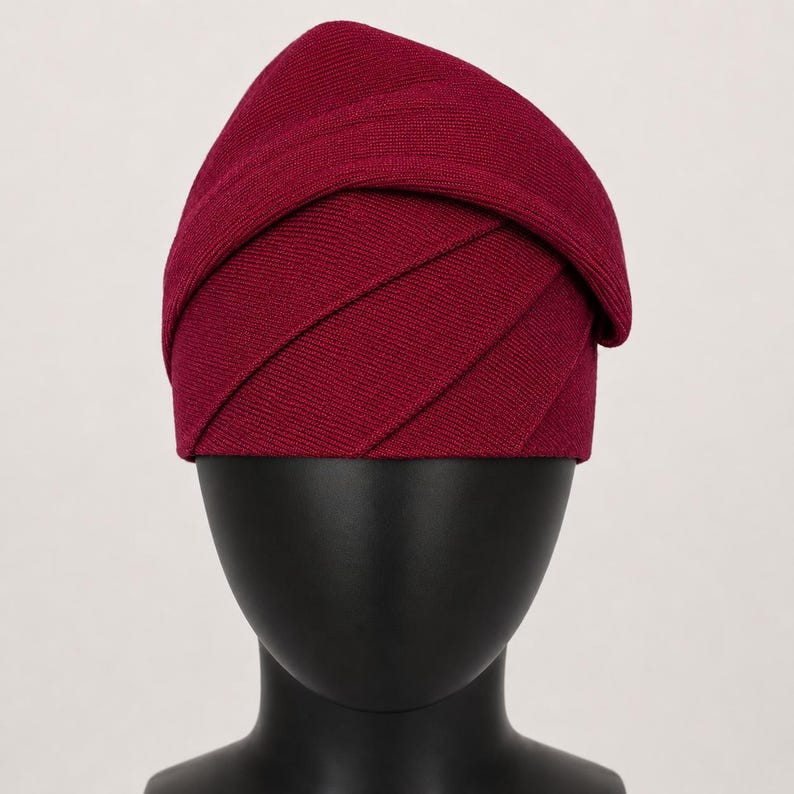 Peut inclure: Un chapeau bordeaux au design superpos&eacute; unique. Le chapeau est fait d'un tissu c&ocirc;tel&eacute;, avec des plis horizontaux cr&eacute;ant un aspect textur&eacute;. Il est pr&eacute;sent&eacute; sur une t&ecirc;te de mannequin noire, mettant en valeur sa forme et son style.