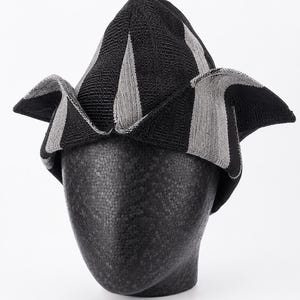 May include: A black and gray striped hat with a unique, folded design. The hat is displayed on a black mannequin head. The fabric has a textured appearance, with alternating black and gray vertical stripes. The hat's brim is folded in a distinctive shape.