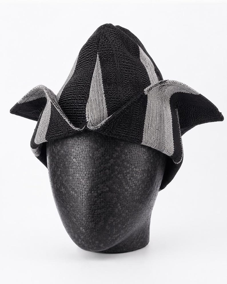 May include: A black and gray striped hat with a unique, folded design. The hat is displayed on a black mannequin head. The fabric has a textured appearance, with alternating black and gray vertical stripes. The hat's brim is folded in a distinctive shape.