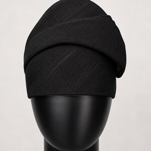 Peut inclure: Un chapeau de style turban noir en tissu textur&eacute;. Le chapeau est pr&eacute;sent&eacute; sur une t&ecirc;te de mannequin noire. Le design comprend une bande envelopp&eacute;e sur le dessus, cr&eacute;ant un look &eacute;l&eacute;gant. Le chapeau est de couleur noire unie.