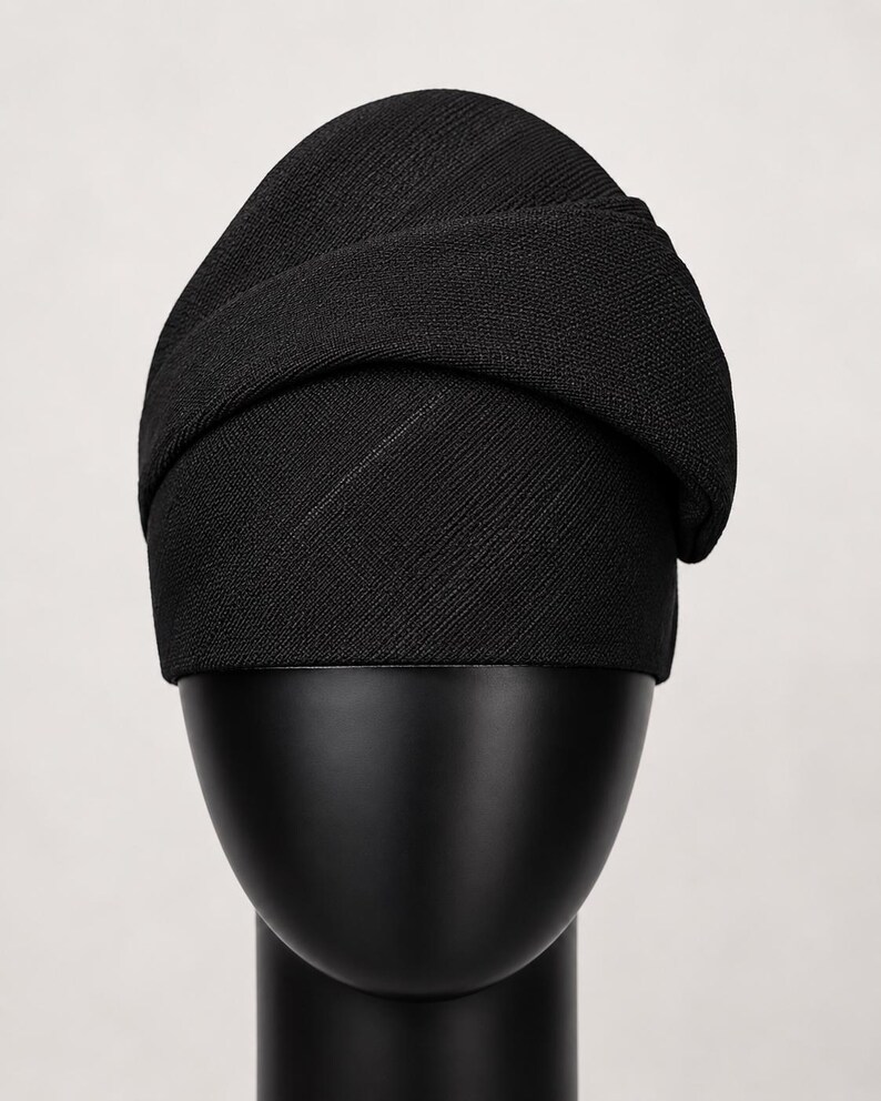 Peut inclure: Un chapeau de style turban noir en tissu textur&eacute;. Le chapeau est pr&eacute;sent&eacute; sur une t&ecirc;te de mannequin noire. Le design comprend une bande envelopp&eacute;e sur le dessus, cr&eacute;ant un look &eacute;l&eacute;gant. Le chapeau est de couleur noire unie.