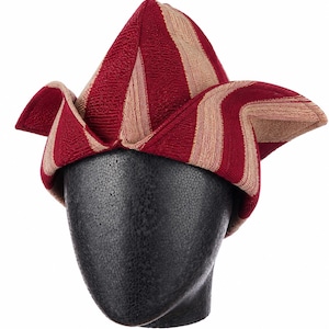 May include: A red and tan striped hat with a unique, folded design. The hat is displayed on a black mannequin head. The hat's fabric has a woven texture, and the stripes are vertical on the pointed crown.