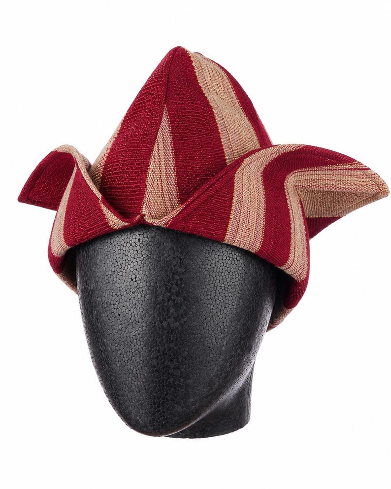 May include: A red and tan striped hat with a unique, folded design. The hat is displayed on a black mannequin head. The hat's fabric has a woven texture, and the stripes are vertical on the pointed crown.