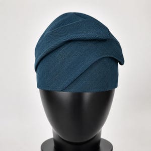 Peut inclure: Un chapeau de couleur sarcelle avec un design superpos&eacute;. Le chapeau est fait d'un tissu textur&eacute; et repose sur une t&ecirc;te de mannequin noire. Le design du chapeau pr&eacute;sente des panneaux de tissu qui se chevauchent, cr&eacute;ant une apparence unique et &eacute;l&eacute;gante. C'est un accessoire de mode.
