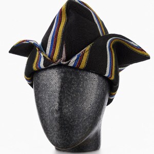 May include: A black hat with colorful stripes of yellow, red, blue, and white. The hat is displayed on a black mannequin head. The hat's design features a folded, peaked crown and flared sides, showcasing a vibrant, patterned fabric.