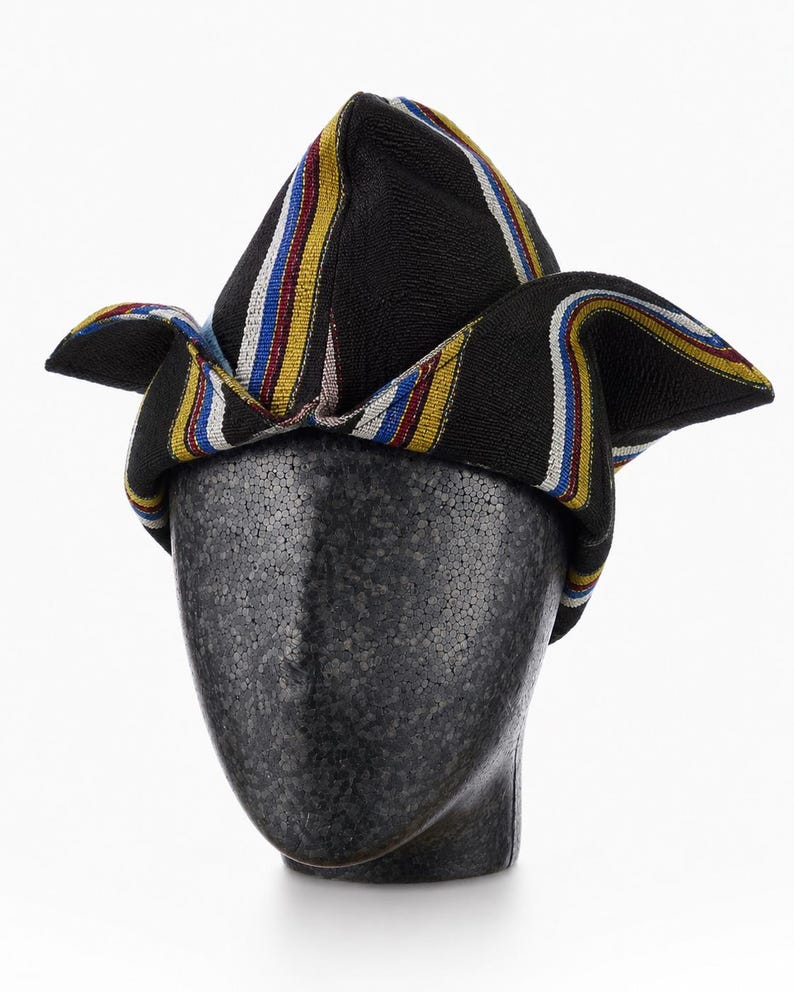 May include: A black hat with colorful stripes of yellow, red, blue, and white. The hat is displayed on a black mannequin head. The hat's design features a folded, peaked crown and flared sides, showcasing a vibrant, patterned fabric.