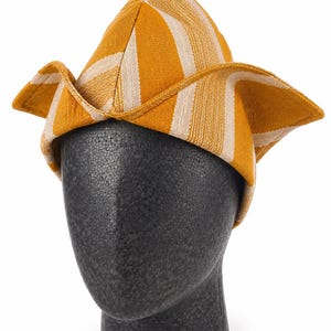 May include: A yellow and white striped hat with a unique folded brim. The hat is displayed on a black mannequin head. The hat's design features a combination of yellow and white vertical stripes, creating a visually appealing pattern. The brim is folded up on the sides.