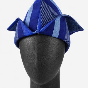 May include: A blue and white striped hat with a unique folded design. The hat is displayed on a black mannequin head. The hat's color scheme includes shades of blue and white, creating a visually appealing accessory.