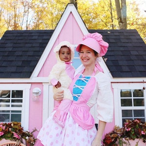 May include: A woman in a pink and white dress and a pink and white hat holds a baby dressed as a lamb in front of a pink playhouse.