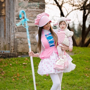 May include: A woman dressed as Bo Peep from Toy Story is holding a child dressed as a sheep. The woman is wearing a pink dress with white polka dots, a blue and white apron, and a pink hat. She is holding a blue and white candy cane stick. The child is wearing a white sheep costume with pink accents. They are standing in a grassy area.