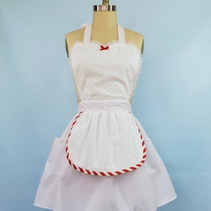 May include: White apron with a red and white striped trim around the bottom edge. The apron has a heart-shaped top with a red bow and a gathered skirt.