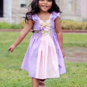May include: A young girl wearing a lavender and pink princess dress with a gold bodice and a floral crown. The dress has a layered skirt and is paired with pink shoes. The girl is smiling and standing on green grass.