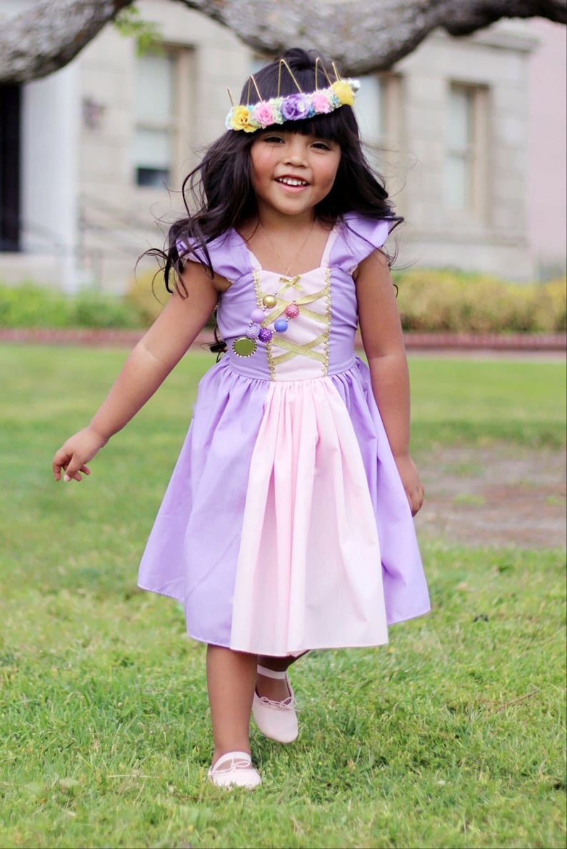 May include: A young girl wearing a lavender and pink princess dress with a gold bodice and a floral crown. The dress has a layered skirt and is paired with pink shoes. The girl is smiling and standing on green grass.