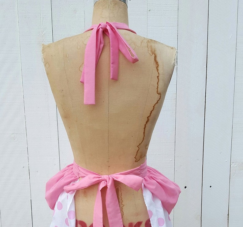 May include: Pink and white polka dot apron with a ruffled hem and pink ties. The apron is tied around a dress form.