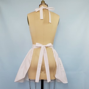 May include: White apron with a gathered skirt and two pockets. The apron ties at the neck and waist with white ribbon.
