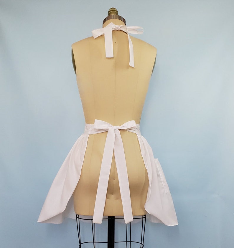 May include: White apron with a gathered skirt and two pockets. The apron ties at the neck and waist with white ribbon.