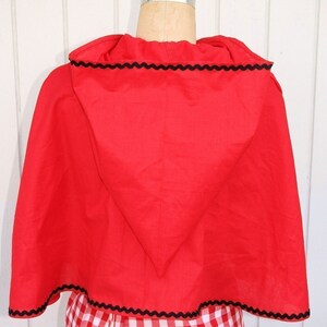 May include: Red hooded cape with black scalloped trim. The cape is made of a soft, red fabric and has a pointed hood. The cape is worn over a red and white gingham dress.
