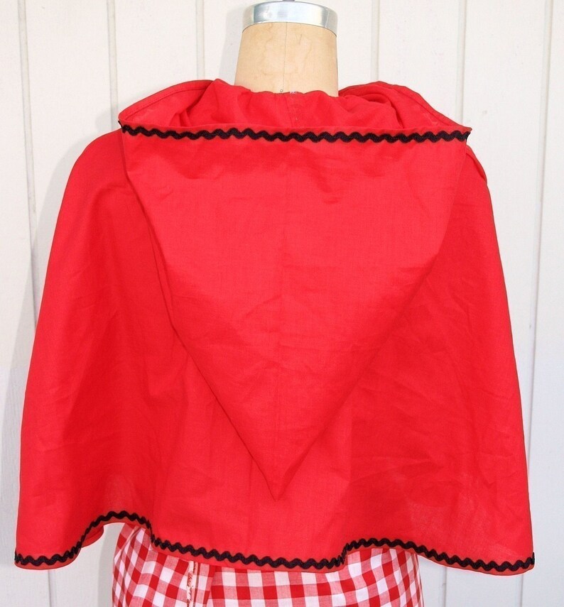 May include: Red hooded cape with black scalloped trim. The cape is made of a soft, red fabric and has a pointed hood. The cape is worn over a red and white gingham dress.