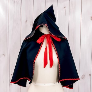 May include: A black hooded cape with red trim and a red ribbon tied at the neck. The cape is made of a soft, flowing fabric and has a classic, elegant design.