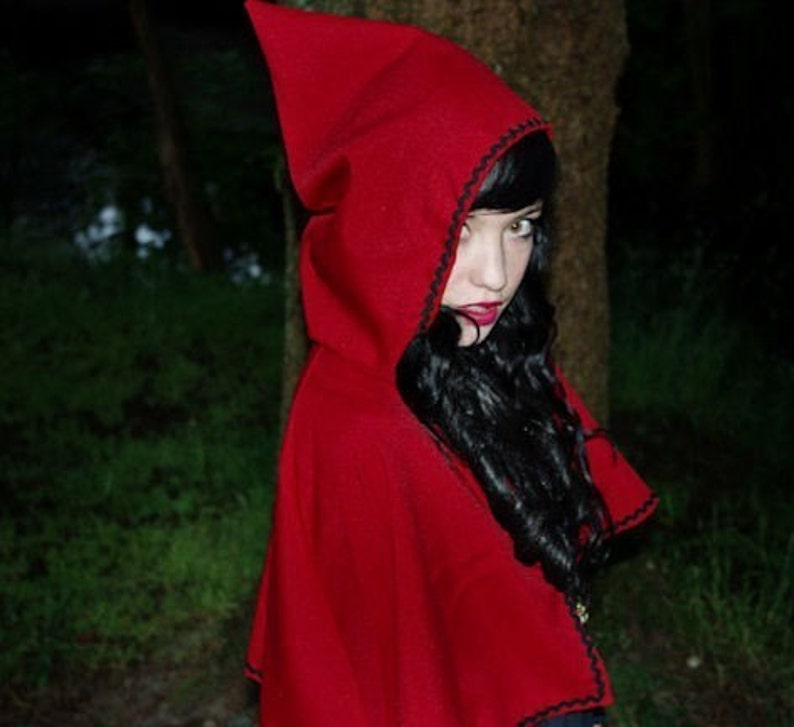 May include: A woman wearing a red hooded cape with black trim. The hood is pulled up over her head, and she is looking at the camera with a serious expression. Her hair is long and black, and she is wearing dark makeup.