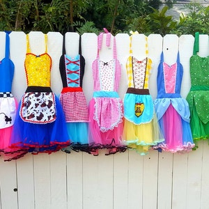 May include: Seven colorful aprons with tulle skirts. The aprons feature different patterns and designs, including a checkered pattern, a cow print, a floral pattern, and a solid color. The aprons are hanging on a white fence.