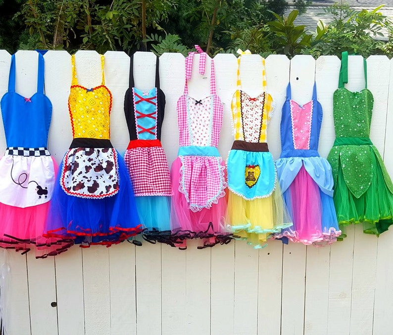 May include: Seven colorful aprons with tulle skirts. The aprons feature different patterns and designs, including a checkered pattern, a cow print, a floral pattern, and a solid color. The aprons are hanging on a white fence.