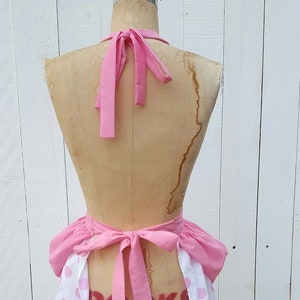 May include: Pink and white polka dot apron with a pink ruffled bottom and pink ties. The apron is tied around a dress form.