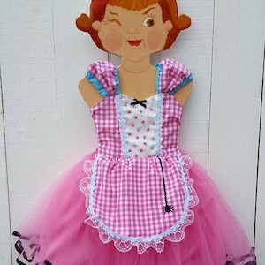 May include: A pink and white gingham dress with a blue and white floral print bodice and a black spider web on the apron. The dress has a tulle skirt and black ribbon trim.