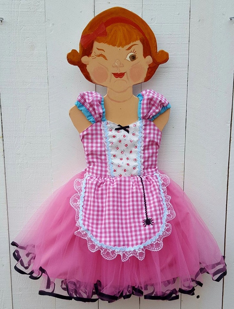 May include: A pink and white gingham dress with a blue and white floral print bodice and a black spider web on the apron. The dress has a tulle skirt and black ribbon trim.