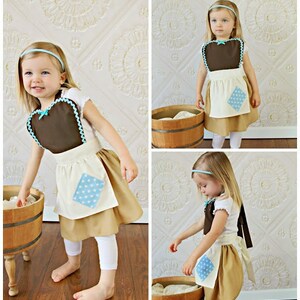 May include: A child's costume featuring a brown bib with a blue bow and trim, paired with a tan skirt and white apron with a blue polka dot patch. The outfit includes a matching blue headband. The costume is designed for a young child.