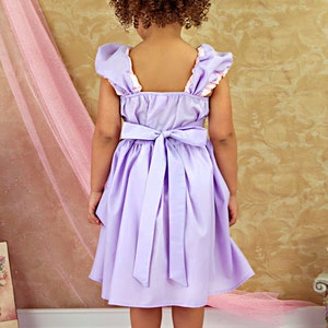 May include: A lavender dress with a bow in the back. The dress has ruffled sleeves with a pink trim. The child is facing away from the camera, with curly brown hair.
