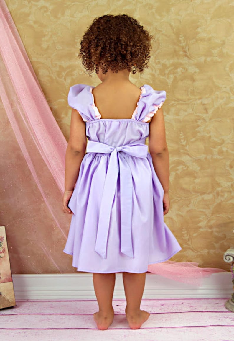 May include: A lavender dress with a bow in the back. The dress has ruffled sleeves with a pink trim. The child is facing away from the camera, with curly brown hair.