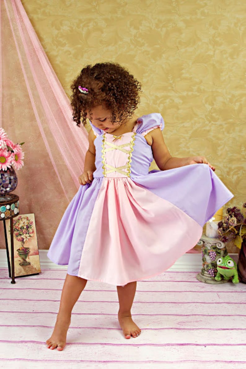 May include: A young girl wearing a princess-style dress in shades of lavender and pink. The dress features a gold-colored bodice with crisscross detailing. The girl is barefoot and standing on a light-colored wooden floor.