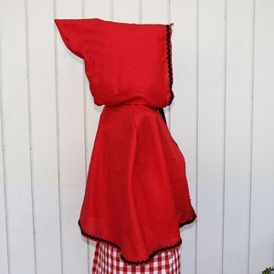 May include: Red hooded cape with black trim. The cape is made of a soft fabric and has a pointed hood.