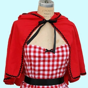 May include: A red hooded cape with black trim and a black ribbon tied at the neck. The cape is worn over a red and white gingham dress with a black belt.