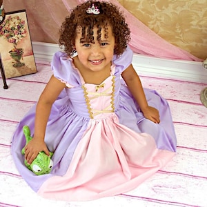 May include: A young child wearing a princess-style dress in shades of lavender and pink, with gold trim and a small tiara. The child is holding a green stuffed animal. The background includes a decorative table and floral arrangements.