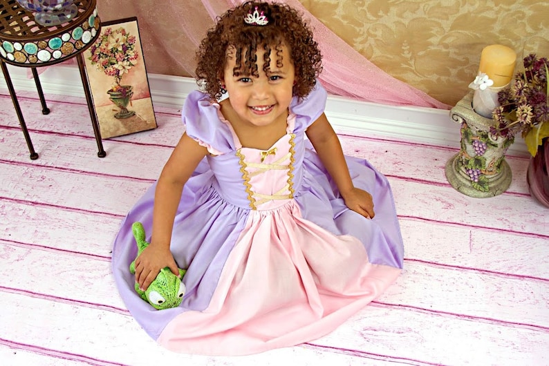 May include: A young child wearing a princess-style dress in shades of lavender and pink, with gold trim and a small tiara. The child is holding a green stuffed animal. The background includes a decorative table and floral arrangements.