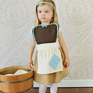 May include: A child's costume featuring a brown bib with blue trim and a white apron with a blue polka-dot patch. The costume includes a tan skirt, white leggings, and a blue headband. A wooden tub is in the background.