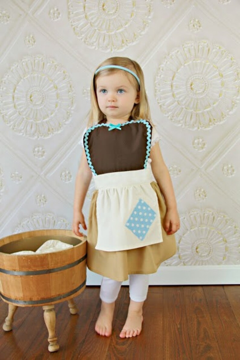 May include: A child's costume featuring a brown bib with blue trim and a white apron with a blue polka-dot patch. The costume includes a tan skirt, white leggings, and a blue headband. A wooden tub is in the background.