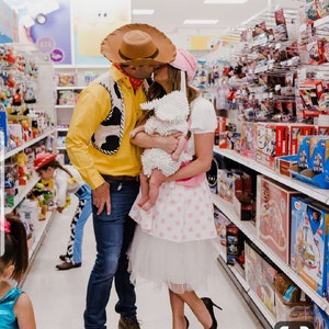 May include: A couple dressed as Woody and Jessie from Toy Story, holding a baby dressed as a sheep, in a toy aisle of a store. The couple is kissing, and the baby is looking at the camera.