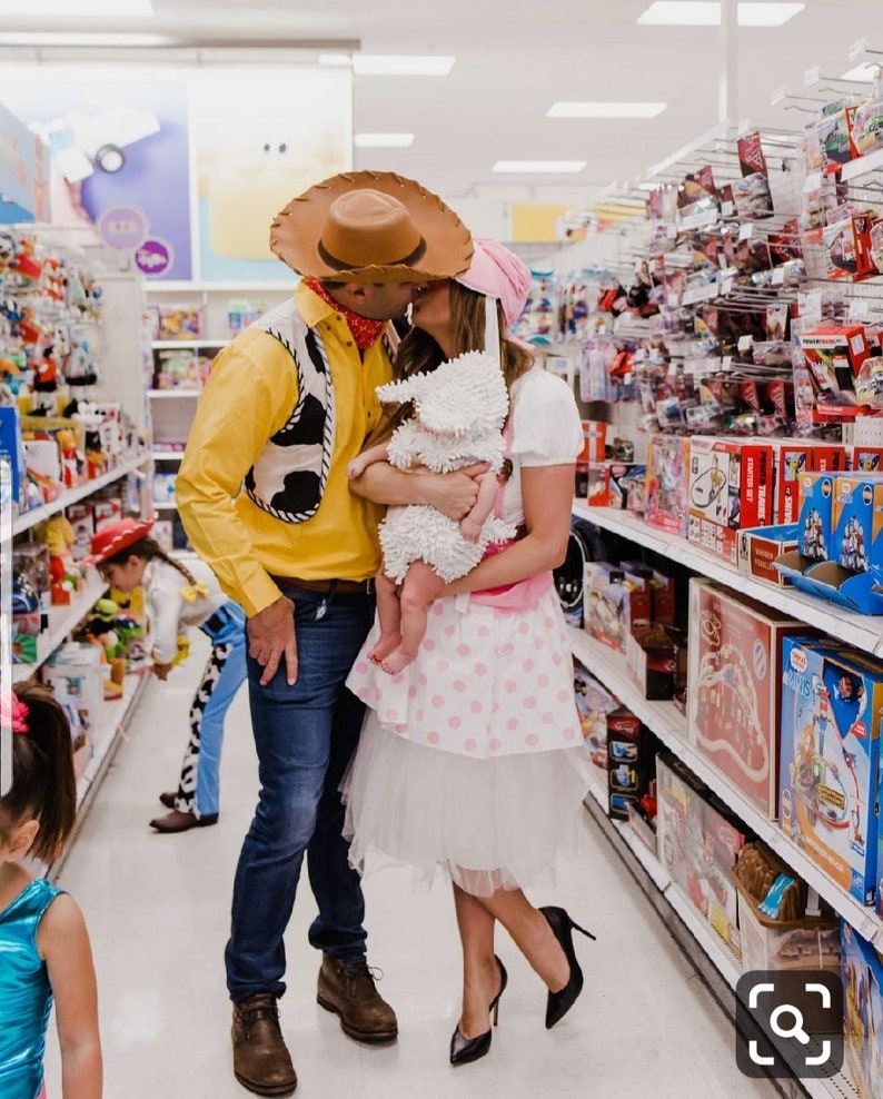 May include: A couple dressed as Woody and Jessie from Toy Story, holding a baby dressed as a sheep, in a toy aisle of a store. The couple is kissing, and the baby is looking at the camera.