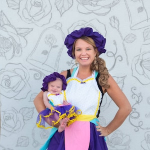 May include: A woman and a baby girl wearing matching purple and yellow dresses and hats. The dresses have a white apron with lace trim and a pink pocket. The woman is holding the baby in her arms. The background is a white wall with a pattern of roses.