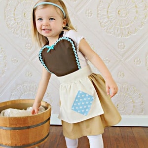 May include: A child's costume featuring a brown apron with a blue scalloped trim and a matching bow. The costume includes a tan skirt, white leggings, and a blue headband. A wooden wash tub is in the foreground.