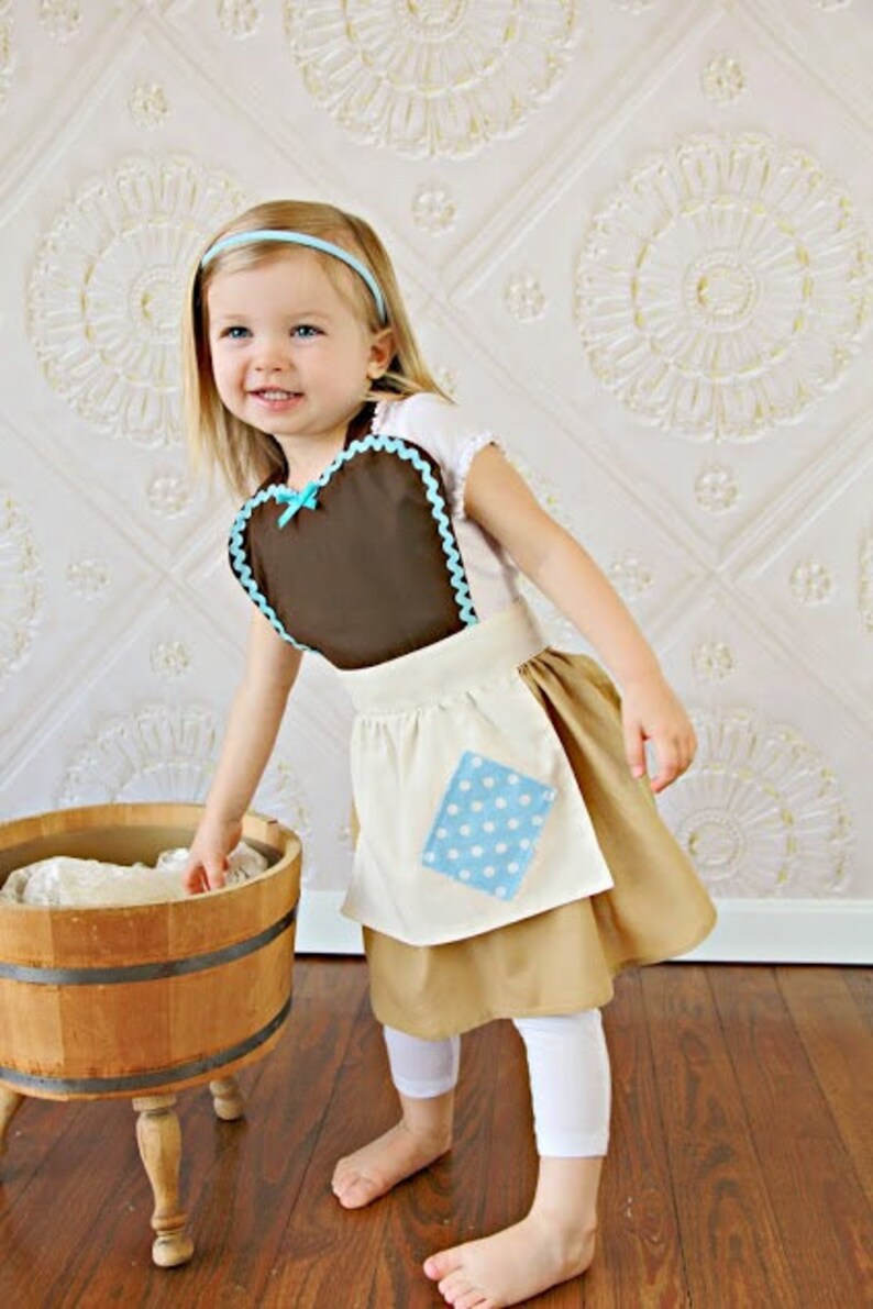 May include: A child's costume featuring a brown apron with a blue scalloped trim and a matching bow. The costume includes a tan skirt, white leggings, and a blue headband. A wooden wash tub is in the foreground.