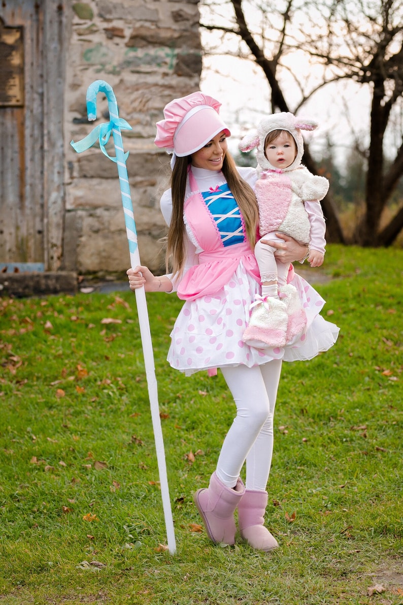May include: A woman dressed as Bo Peep from Toy Story is holding a child dressed as a sheep. The woman is wearing a pink dress with white polka dots, a blue and white apron, and a pink hat. She is holding a blue and white candy cane stick. The child is wearing a white sheep costume with pink accents. They are standing in a grassy area.