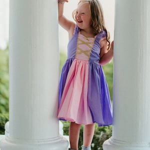May include: A young girl wearing a purple, pink, and gold dress, standing between two white pillars. The dress has a bodice with criss-cross gold detailing and a layered skirt. She is smiling and wearing silver shoes.